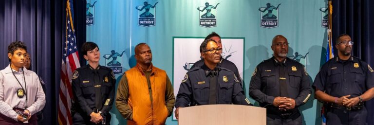 The Detroit Chief of Police stands at a podium with six police officers standing behind. There is a backdrop with the City of Detroit logo and most of the police officers are in uniform including the chief of police.